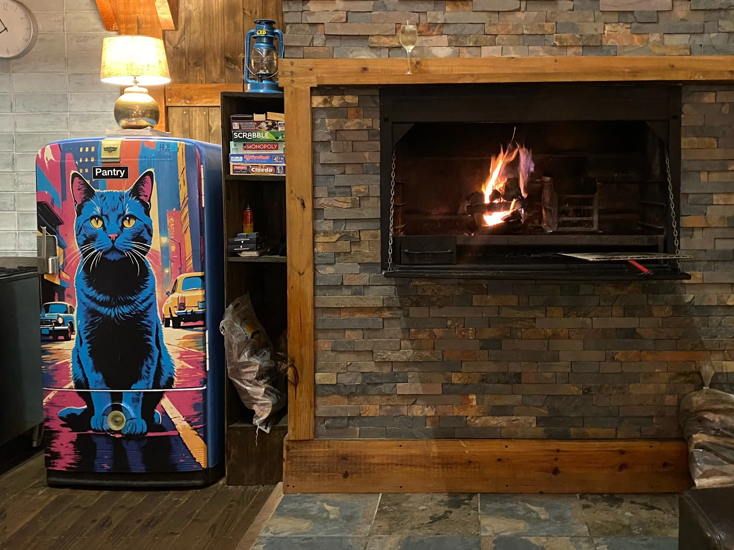 A fire burning in the stone-built indoor braai, with a glass of wine on the timber mantel and board games on the shelf beside it