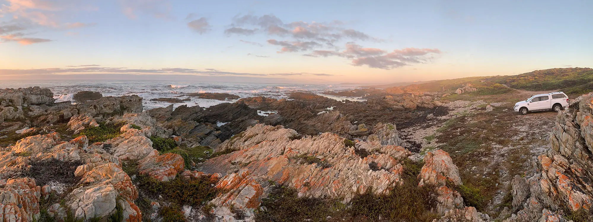 Low sun on orange-lichened rock and coastal fynbos, surf breaking beyond, with a white bakkie parked on the track at the top right